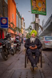 Sikh man seated in the foreground behind him rows of shophouses in vivid colour and detail on a street in Bombay