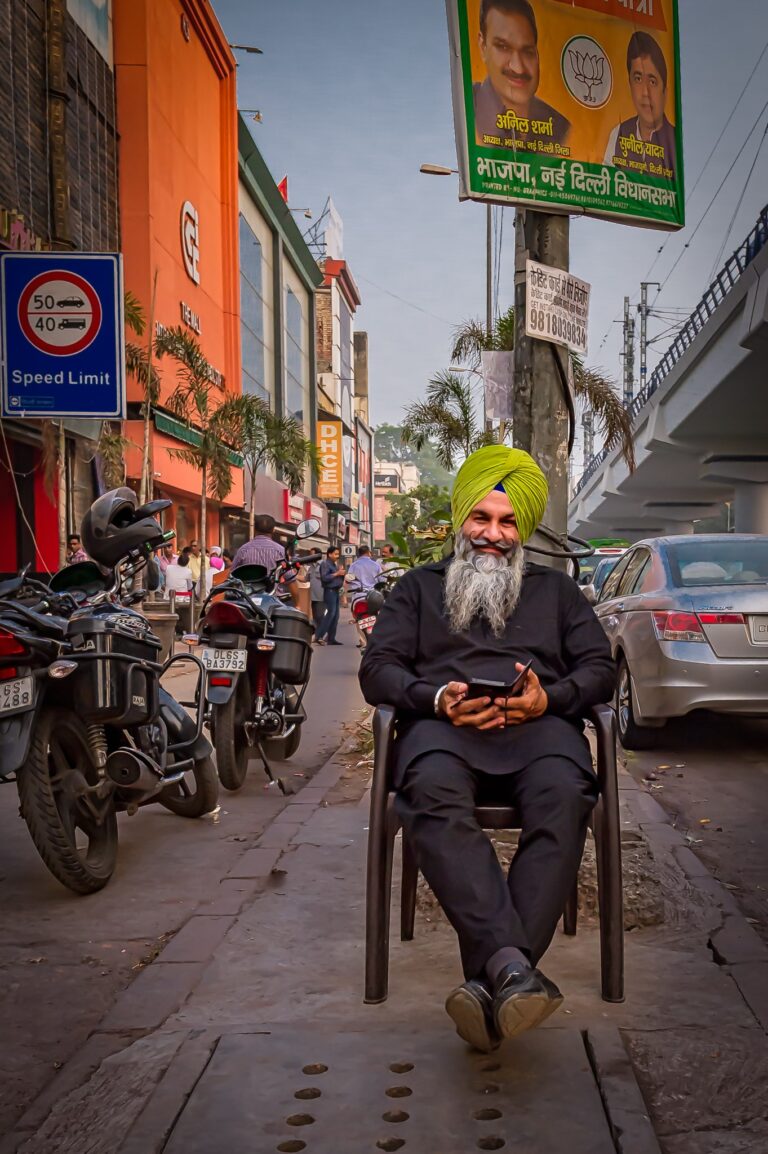 Sikh man seated in the foreground behind him rows of shophouses in vivid colour and detail on a street in Bombay