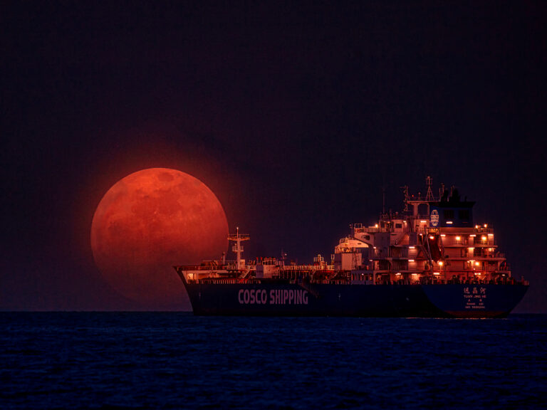 red moon setting behind a large tanker ship